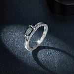 CARISSIE radiant moissanite bezel ring, angled view showing the teal-green stone and smooth silver-tone band on a glittering surface