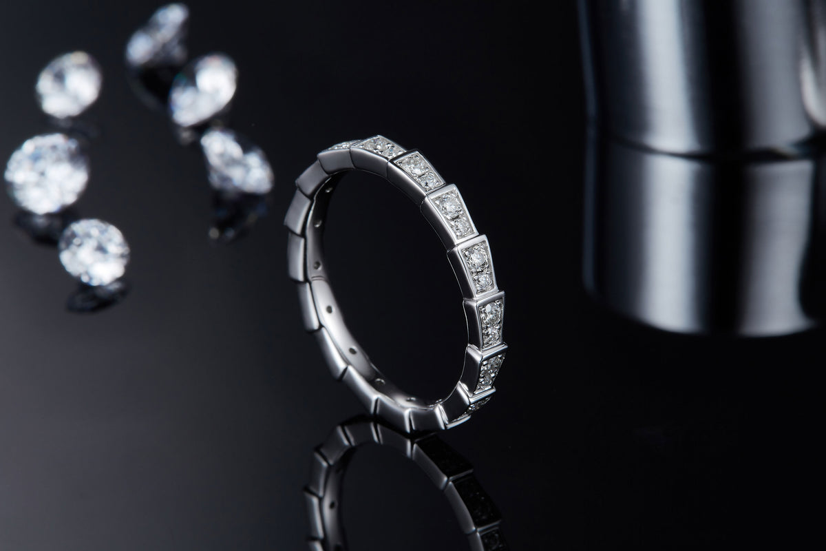 CARISSIE round moissanite eternity band, lifestyle shot on dark reflective surface surrounded by loose gemstones emphasizing brilliance and contrast