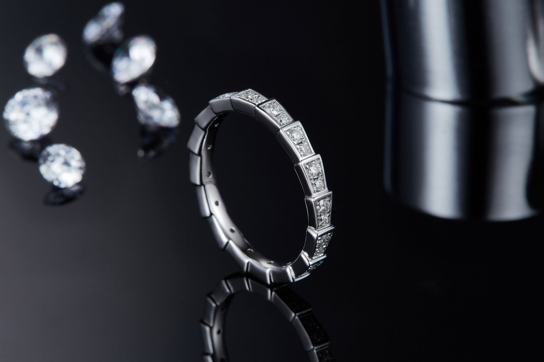 CARISSIE round moissanite eternity band, lifestyle shot on dark reflective surface surrounded by loose gemstones emphasizing brilliance and contrast