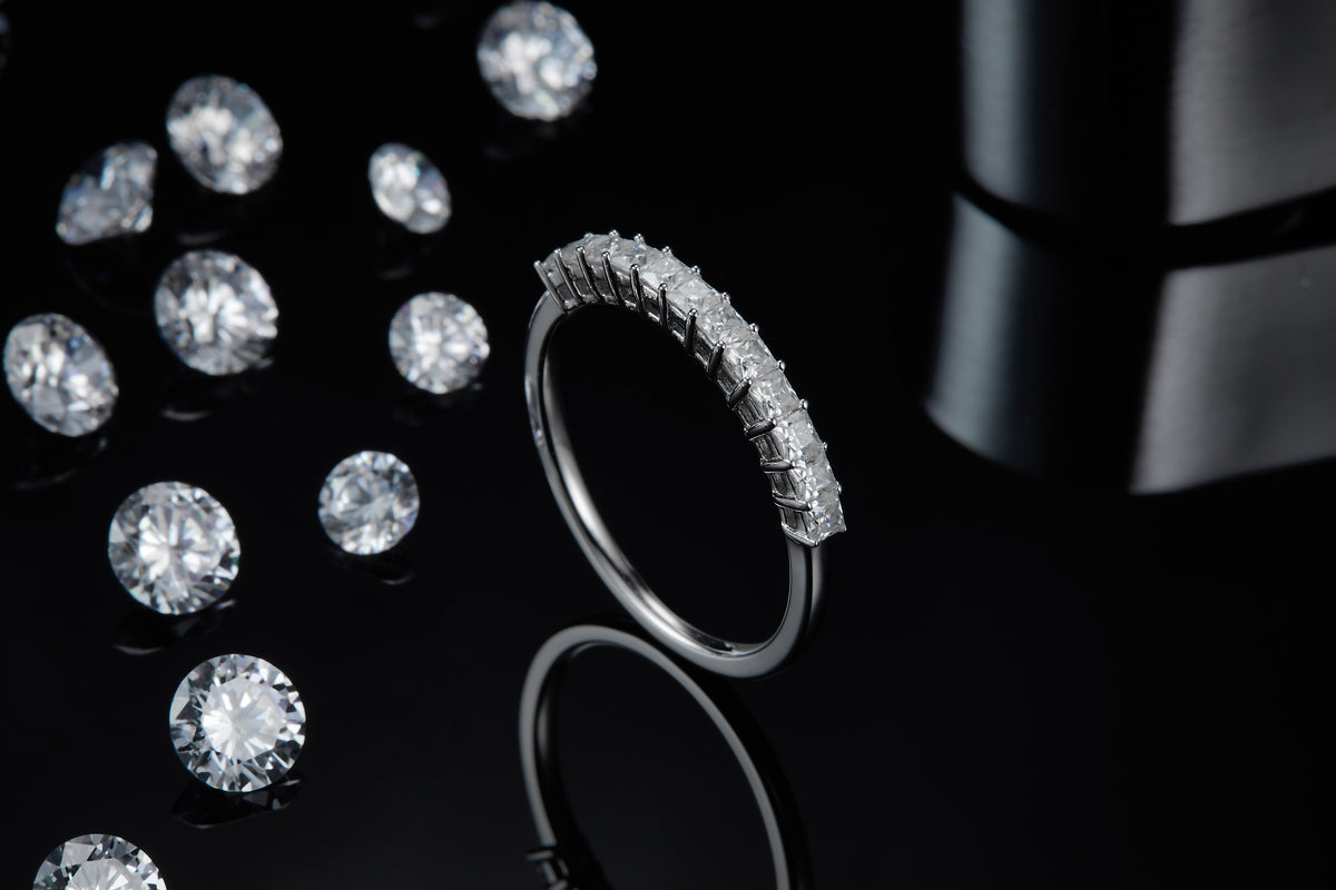 CARISSIE Princess Moissanite Eternity Band, artistic lifestyle shot on a black reflective surface surrounded by loose gemstones to show brilliance.