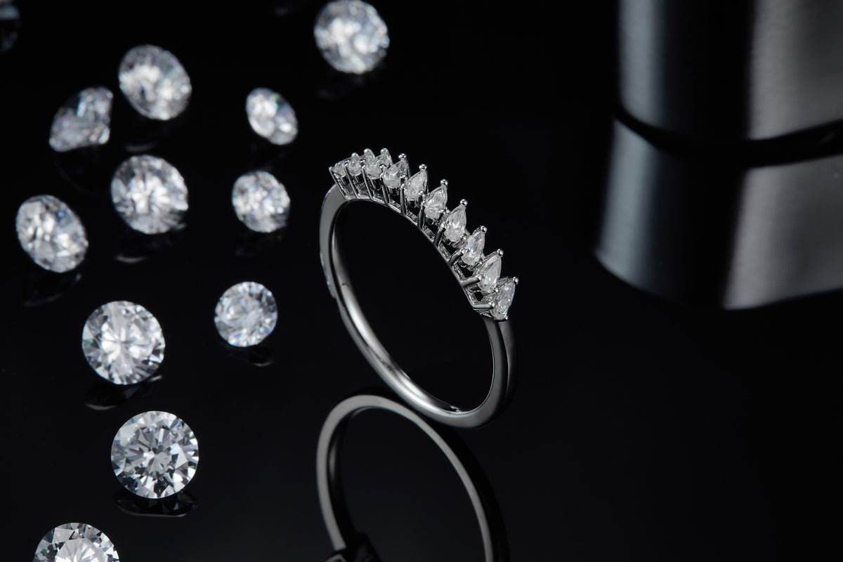 CARISSIE round moissanite solitaire ring, lifestyle shot on a black reflective background with loose gemstones emphasizing sparkle and contrast
