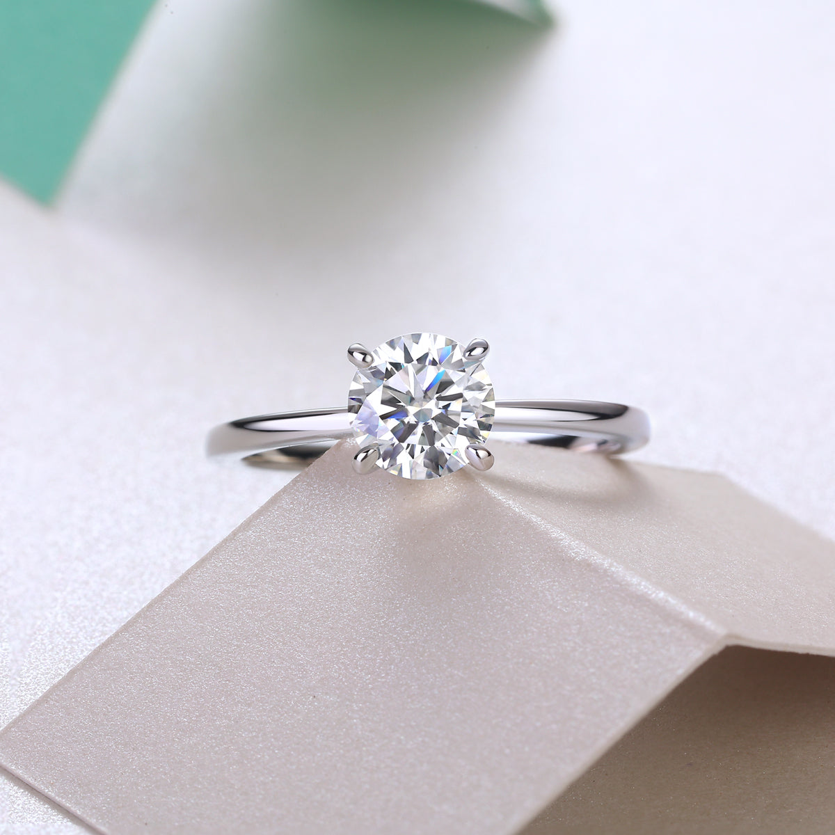 CARISSIE round moissanite solitaire ring resting on textured paper surface, angled to catch light through clear moissanite center stone