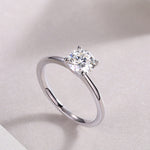 CARISSIE round moissanite solitaire ring diagonal perspective on glittery surface, accentuating reflective band and sparkling gemstone clarity