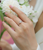 CARISSIE round moissanite solitaire ring on model hand with white flowers background, showing silver-tone metal and brilliant sparkle