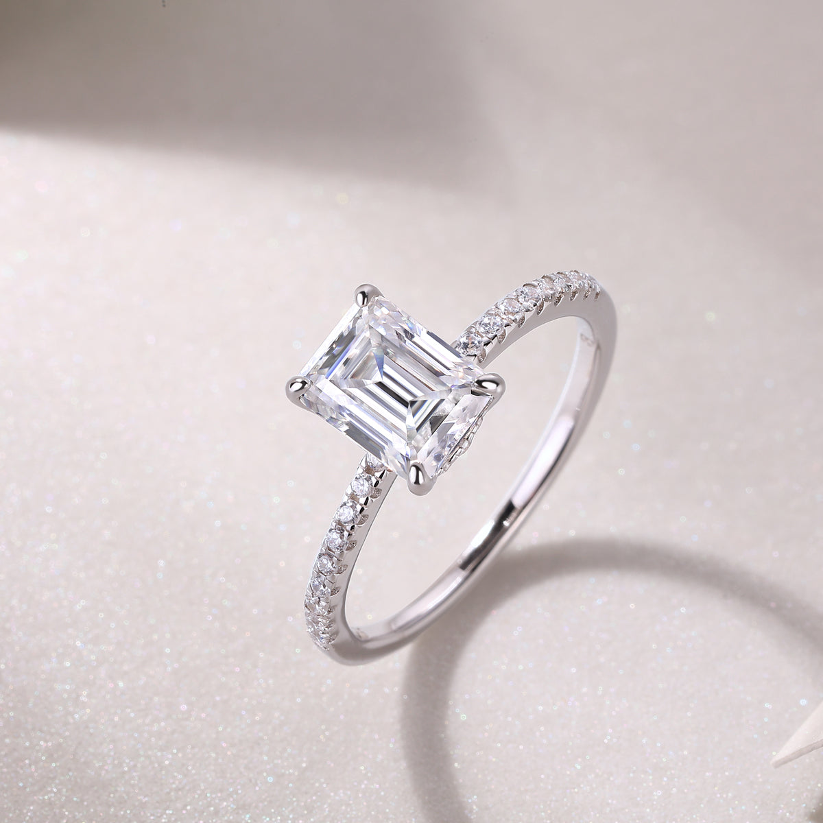 CARISSIE engagement ring diagonal view on glittery background, featuring brilliant emerald cut stone and polished metal