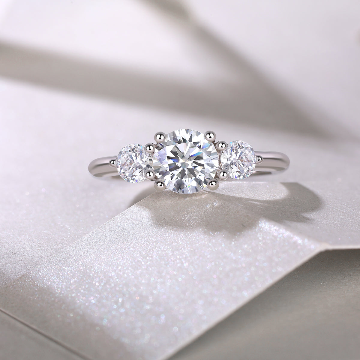 CARISSIE round moissanite solitaire ring, lifestyle shot on textured white surface capturing light reflection in the stones
