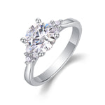 CARISSIE round moissanite solitaire ring, angled view showing six-prong setting and accent stone clusters on silver-tone band