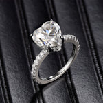 CARISSIE heart solitaire engagement ring, angled view on black leather showing pavé band and four-prong setting