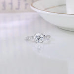 CARISSIE round moissanite solitaire ring, angled view on white surface showing pavé band and center stone sparkle