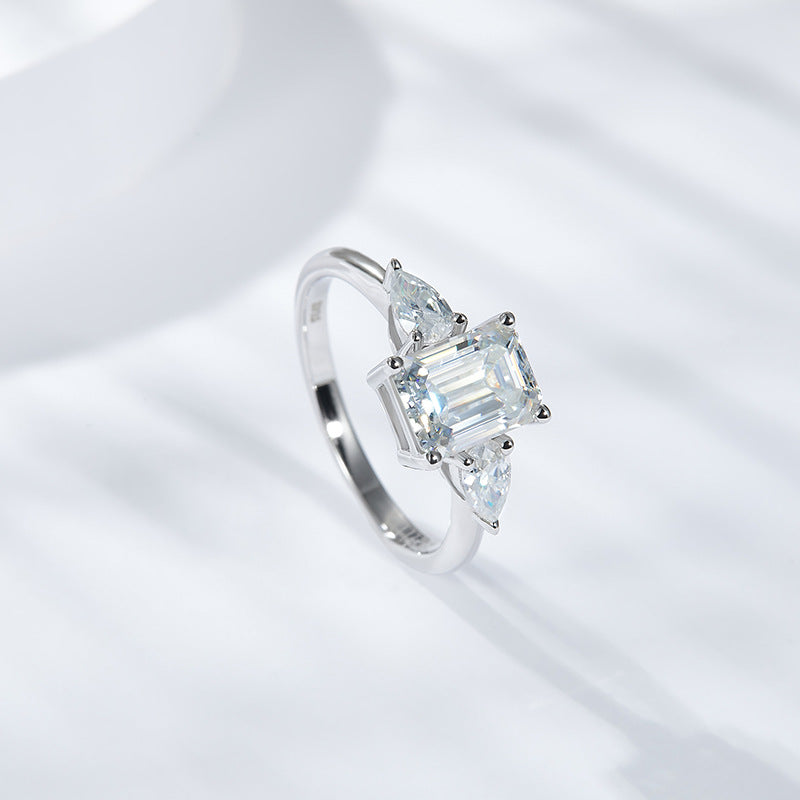 CARISSIE emerald cut solitaire ring, angled view showing pear side stones and white metal band on soft background
