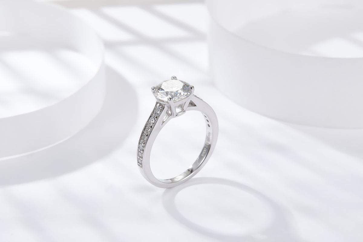 CARISSIE round moissanite solitaire ring, angled lifestyle shot on white surface showcasing high-polish silver finish and pavé details