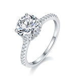 CARISSIE round moissanite solitaire ring, angled view highlighting four-prong setting and pavé diamond band on polished silver-tone metal