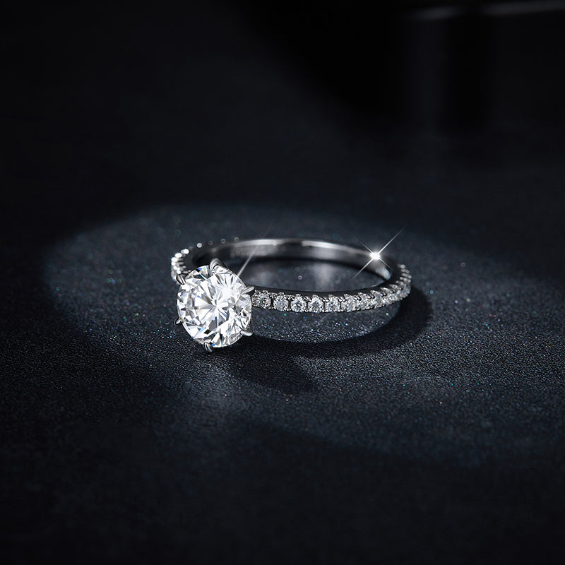 CARISSIE round moissanite solitaire ring, lifestyle angle displaying the half-pavé band design resting on a black textured surface