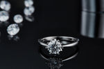 CARISSIE round moissanite solitaire ring, lifestyle shot on a dark reflective surface with scattered loose stones in background