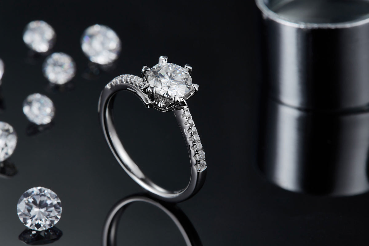 CARISSIE round moissanite solitaire ring, lifestyle shot on black background with loose stones emphasizing the sparkle of the main gem
