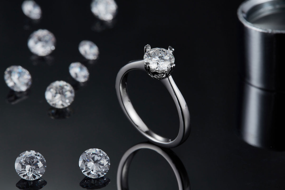 CARISSIE round moissanite solitaire ring, side angle on black surface with scattered loose stones in background
