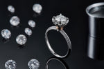 CARISSIE round moissanite halo ring, lifestyle shot on black surface with loose stones showing the ring's profile and sparkle.
