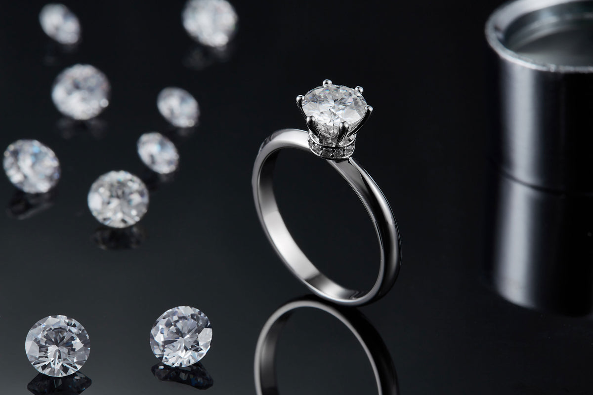 CARISSIE round moissanite solitaire ring, lifestyle shot on black surface with loose stones and reflection