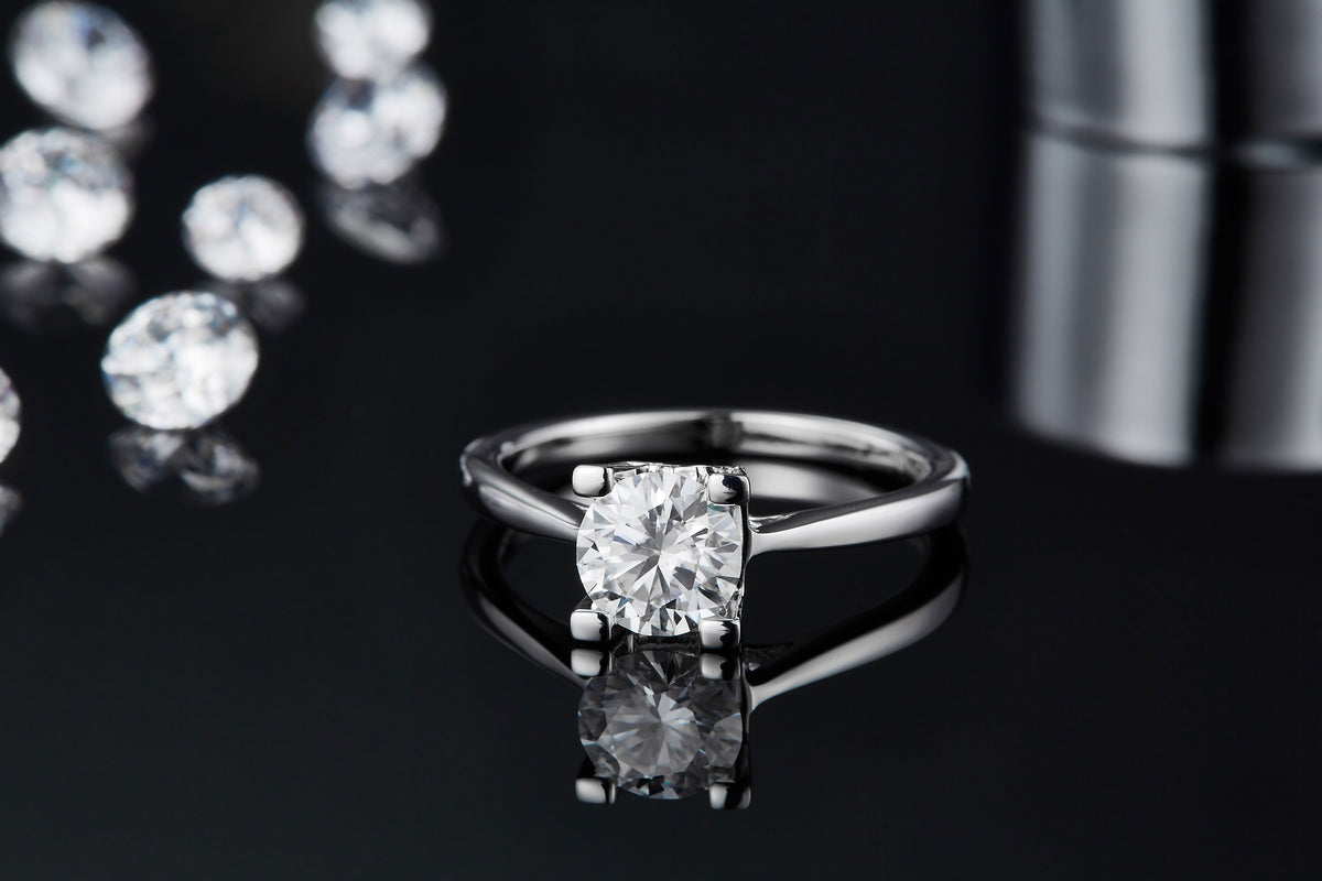CARISSIE round moissanite solitaire ring, low angle view emphasizing the reflection of the round stone and four-prong setting on glossy black