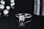 CARISSIE round moissanite solitaire ring, low angle view emphasizing the reflection of the round stone and four-prong setting on glossy black