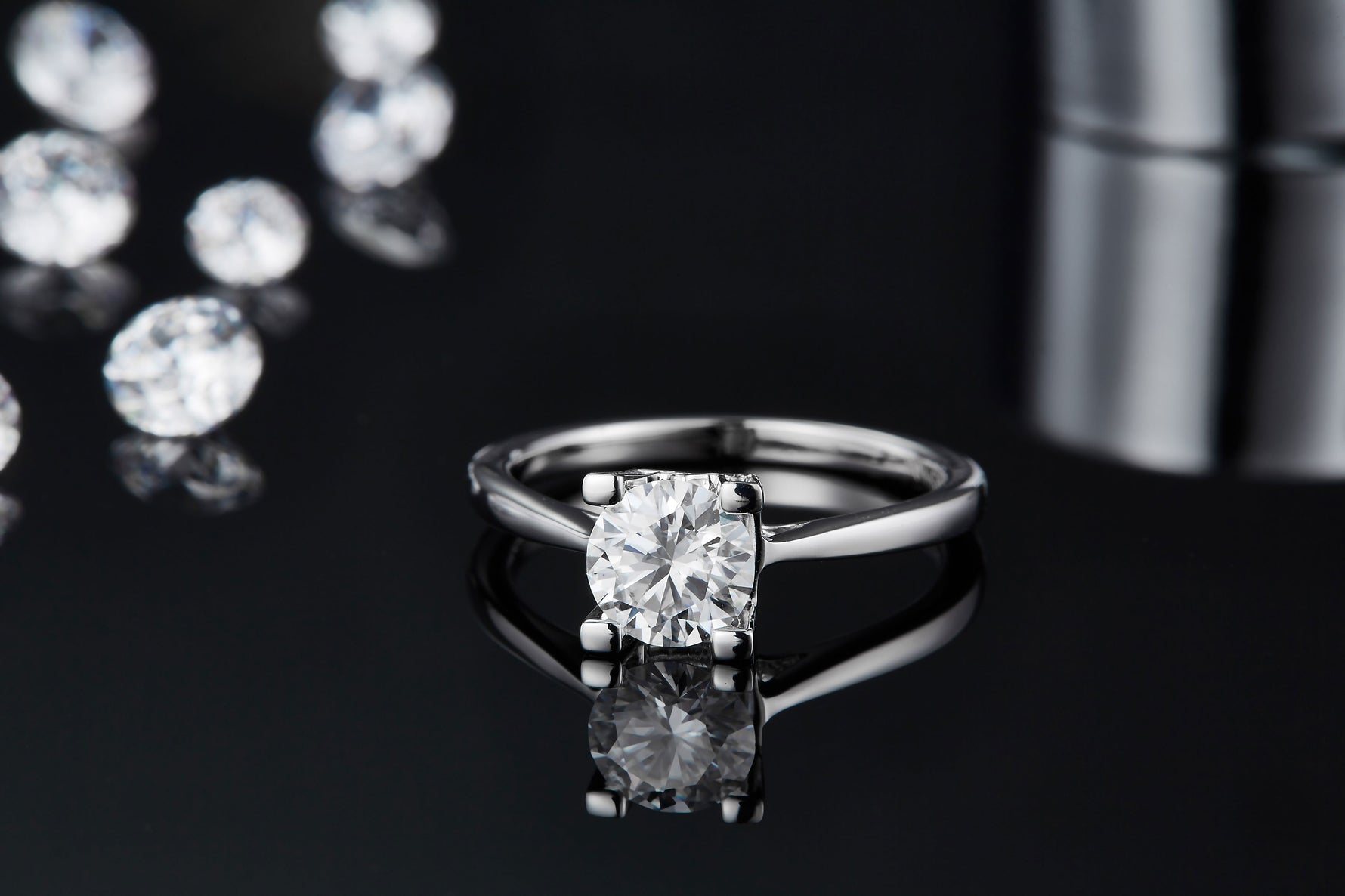 CARISSIE round moissanite solitaire ring, low angle view emphasizing the reflection of the round stone and four-prong setting on glossy black