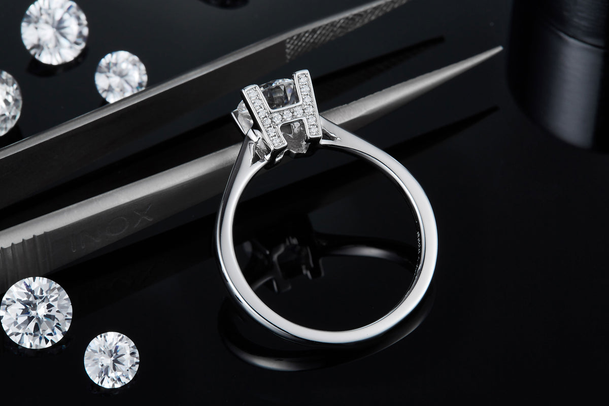 CARISSIE round moissanite solitaire ring, close-up detail of the H-shaped pavé setting held by tweezers next to loose gems