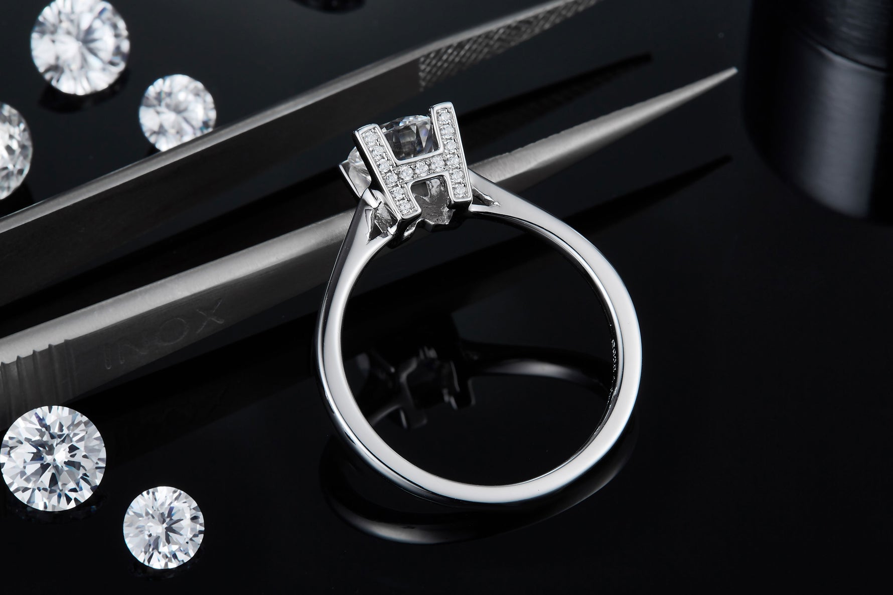 CARISSIE round moissanite solitaire ring, close-up detail of the H-shaped pavé setting held by tweezers next to loose gems