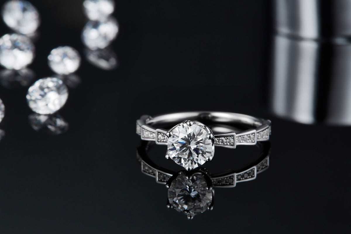 CARISSIE round moissanite solitaire ring, lifestyle shot on reflective surface emphasizing the sparkle of the main stone and side accents