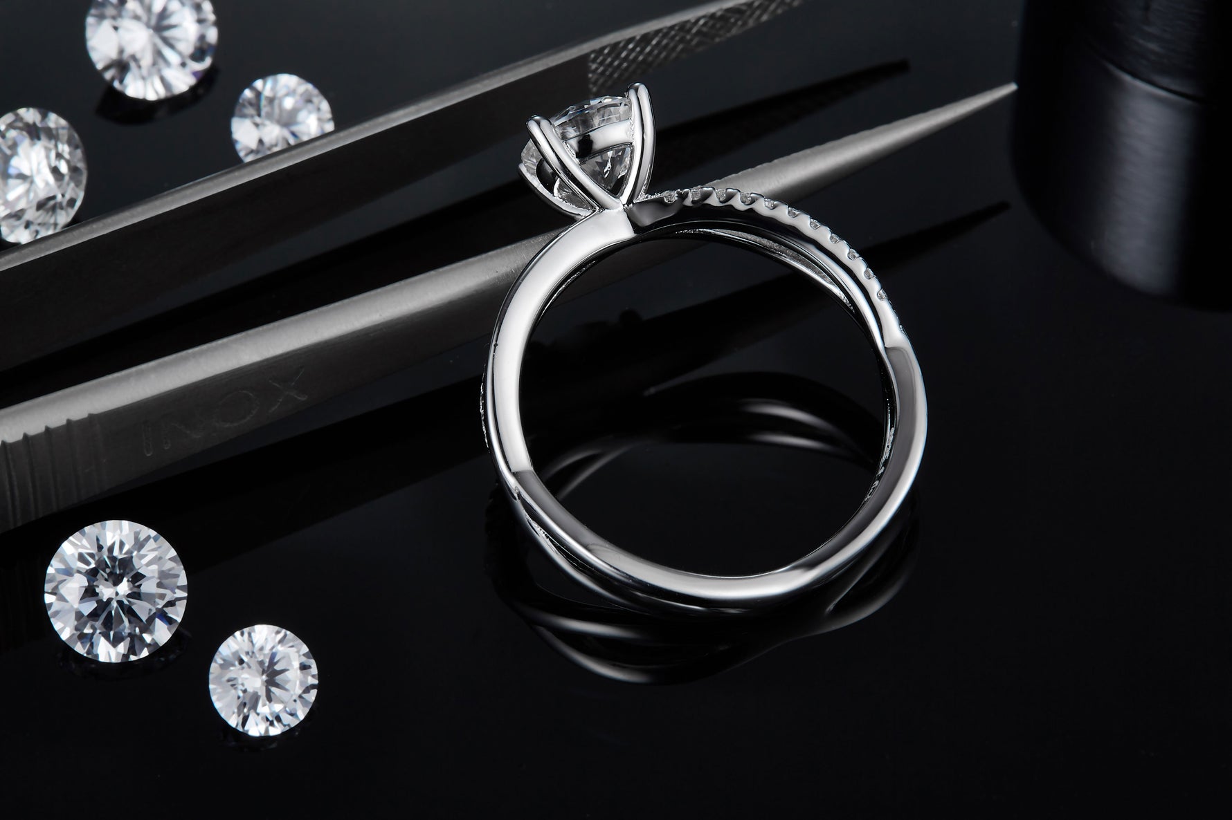 CARISSIE round moissanite solitaire ring, close-up side view resting near tweezers, showcasing the polished metal finish and prong details