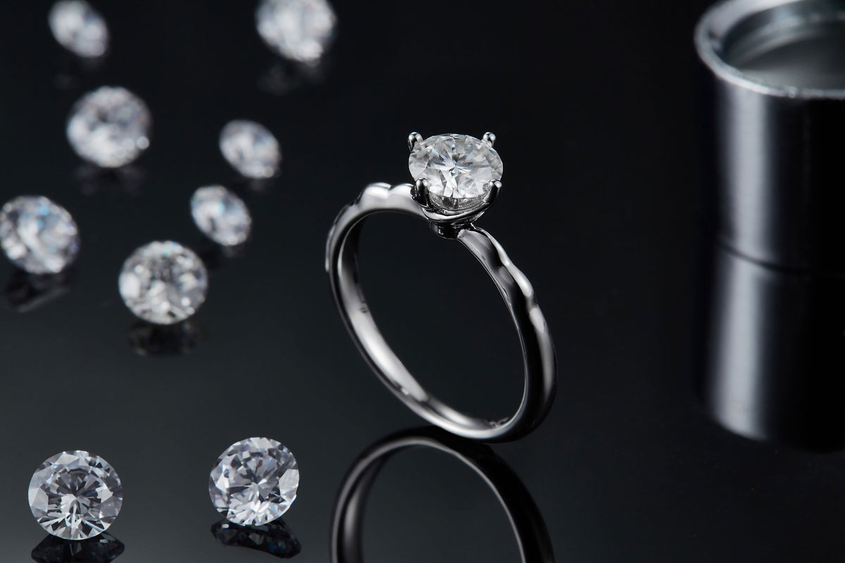 CARISSIE round moissanite solitaire ring, standing upright on a dark reflective surface with loose stones in background