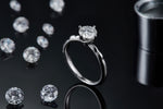 CARISSIE round moissanite solitaire ring, standing upright on a dark reflective surface with loose stones in background