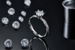 CARISSIE round moissanite solitaire ring, lifestyle shot standing on black surface with loose stones in background