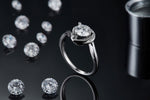 CARISSIE round moissanite engagement ring, lifestyle shot on a black reflective surface with loose gemstones in background