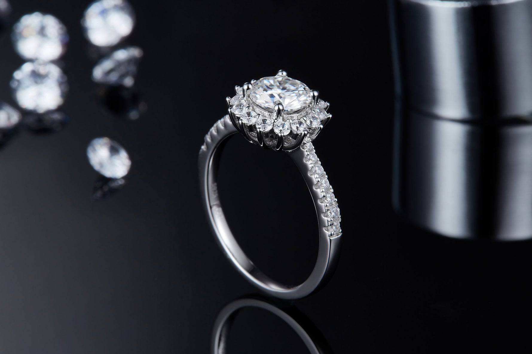 CARISSIE round moissanite halo engagement ring, lifestyle shot on a black surface with loose stones and metallic props.