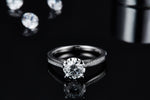 CARISSIE round moissanite solitaire ring, lifestyle shot on black reflective surface with loose stones in background