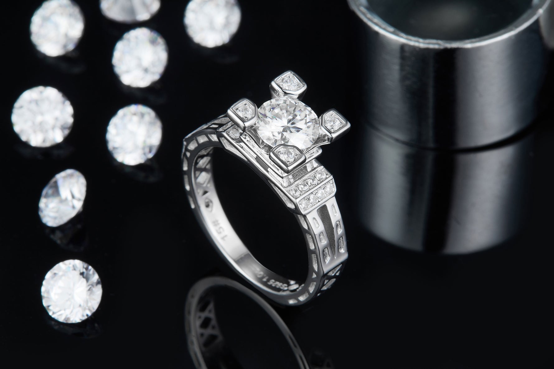 CARISSIE round moissanite solitaire ring, lifestyle shot on black surface displaying sparkle against loose stones and metal cylinder