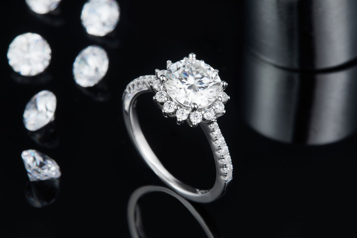 CARISSIE round moissanite halo engagement ring, angled shot on a dark reflective surface with loose gemstones in the background.