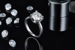 CARISSIE round moissanite solitaire ring, side profile on black background with scattered loose stones and metal cylinder