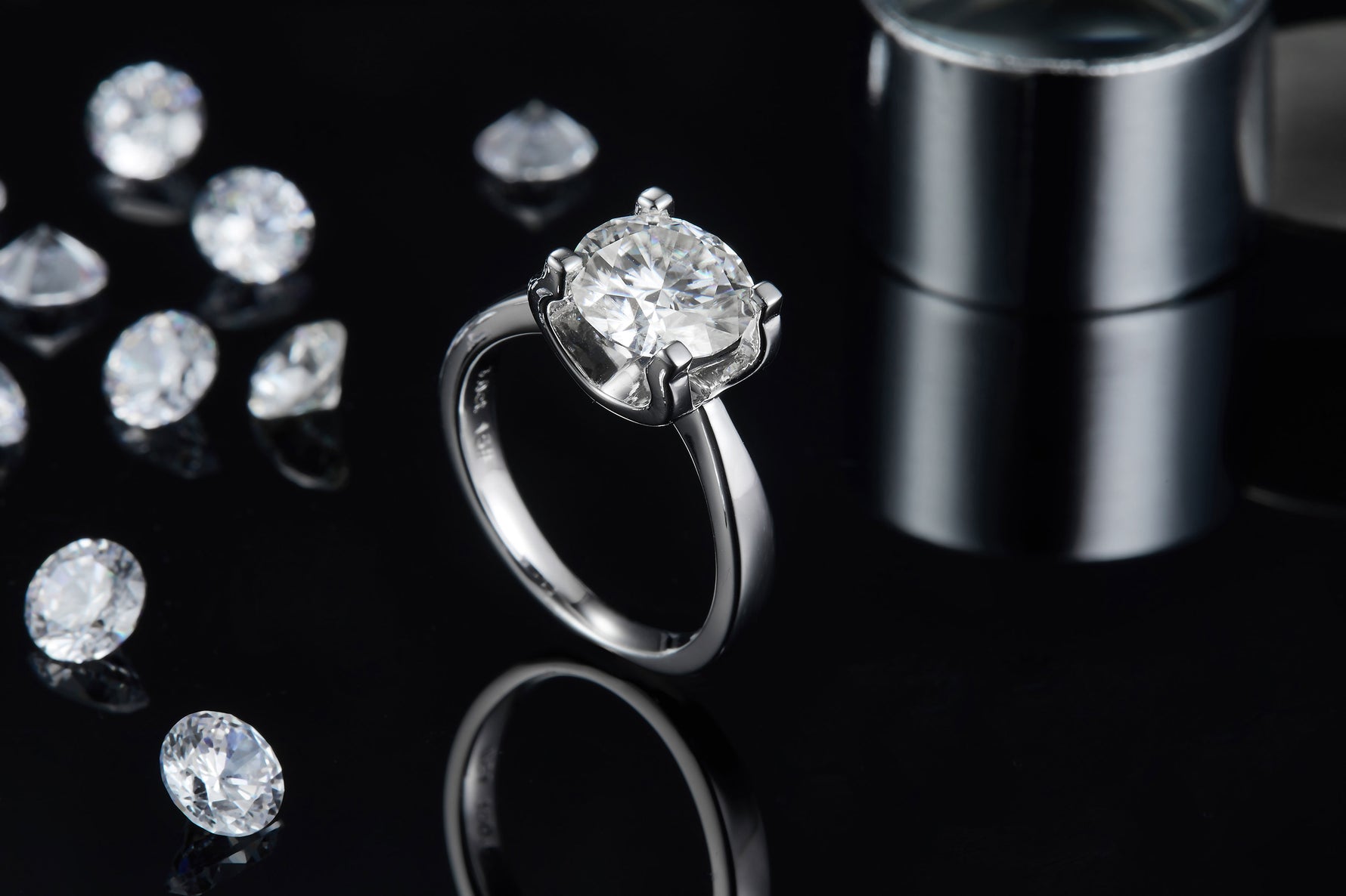 CARISSIE round moissanite solitaire ring, side profile on black background with scattered loose stones and metal cylinder