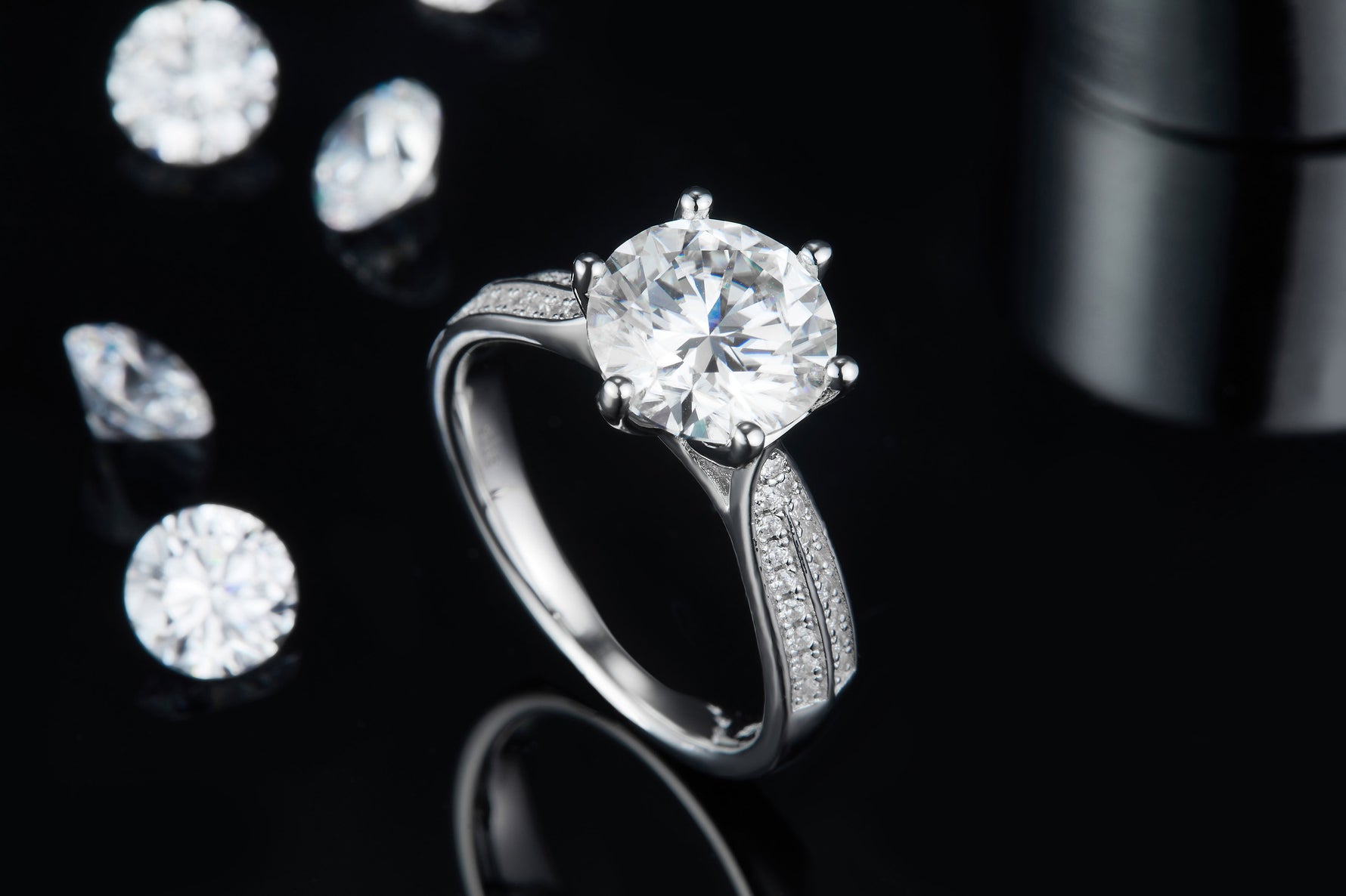 CARISSIE round moissanite solitaire ring, lifestyle shot on black surface displaying brilliance against loose gems