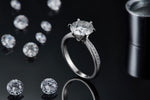 CARISSIE round moissanite solitaire ring, lifestyle shot standing upright on black surface with scattered loose stones in background.