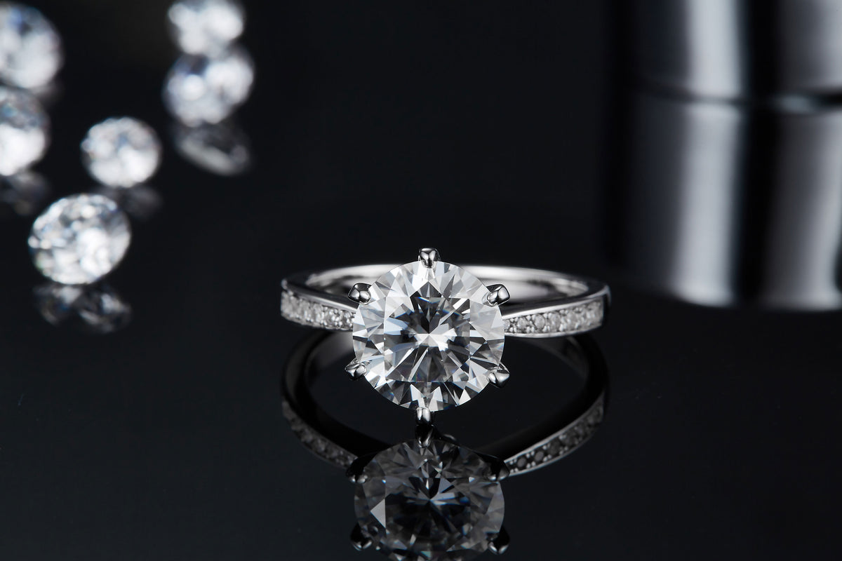 CARISSIE round moissanite solitaire ring, front-facing view resting on reflective black surface showing clear mirror image below.