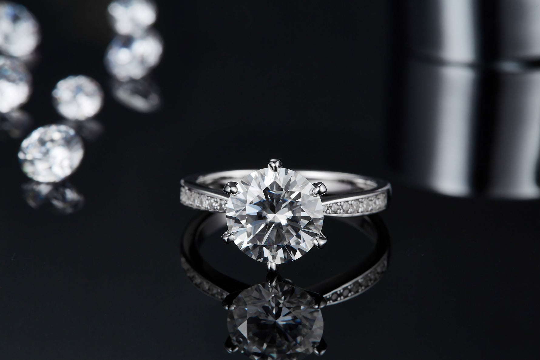 CARISSIE round moissanite solitaire ring, front-facing view resting on reflective black surface showing clear mirror image below.