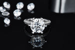 CARISSIE round moissanite ring, lifestyle shot on black reflective surface with loose stones in background