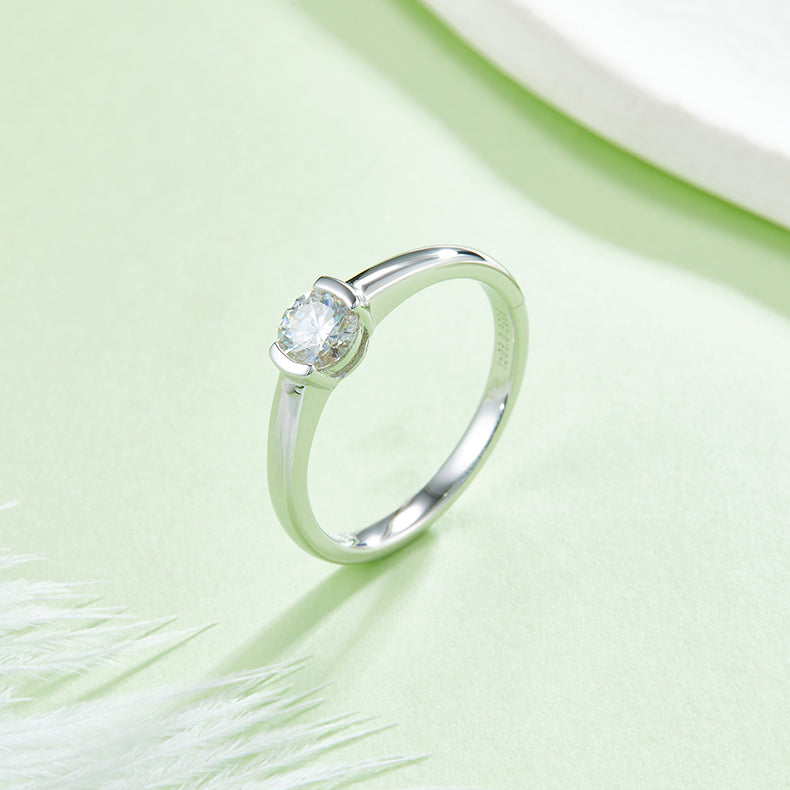CARISSIE round moissanite bezel ring, lifestyle shot on soft green background with white feathers emphasizing the sparkle