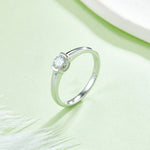 CARISSIE round moissanite bezel ring, lifestyle shot on soft green background with white feathers emphasizing the sparkle