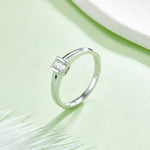 CARISSIE emerald cut bezel engagement ring, lifestyle shot on green background with white feathers highlighting the minimalist design