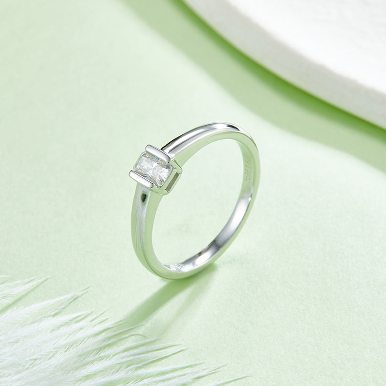 CARISSIE emerald cut bezel engagement ring, lifestyle shot on green background with white feathers highlighting the minimalist design