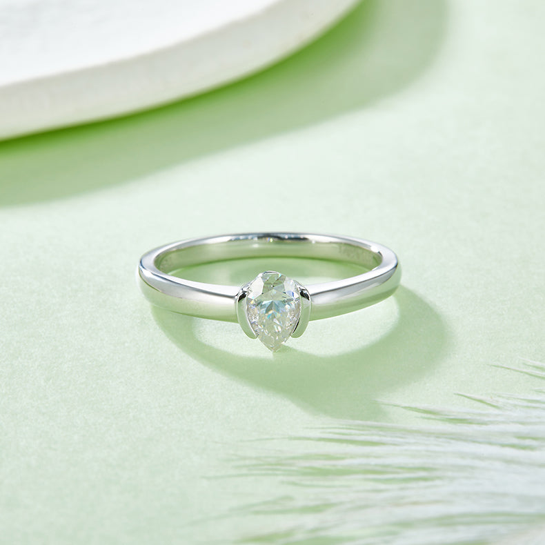 CARISSIE pear moissanite bezel engagement ring, lifestyle shot on soft green surface displaying the teardrop gemstone shape