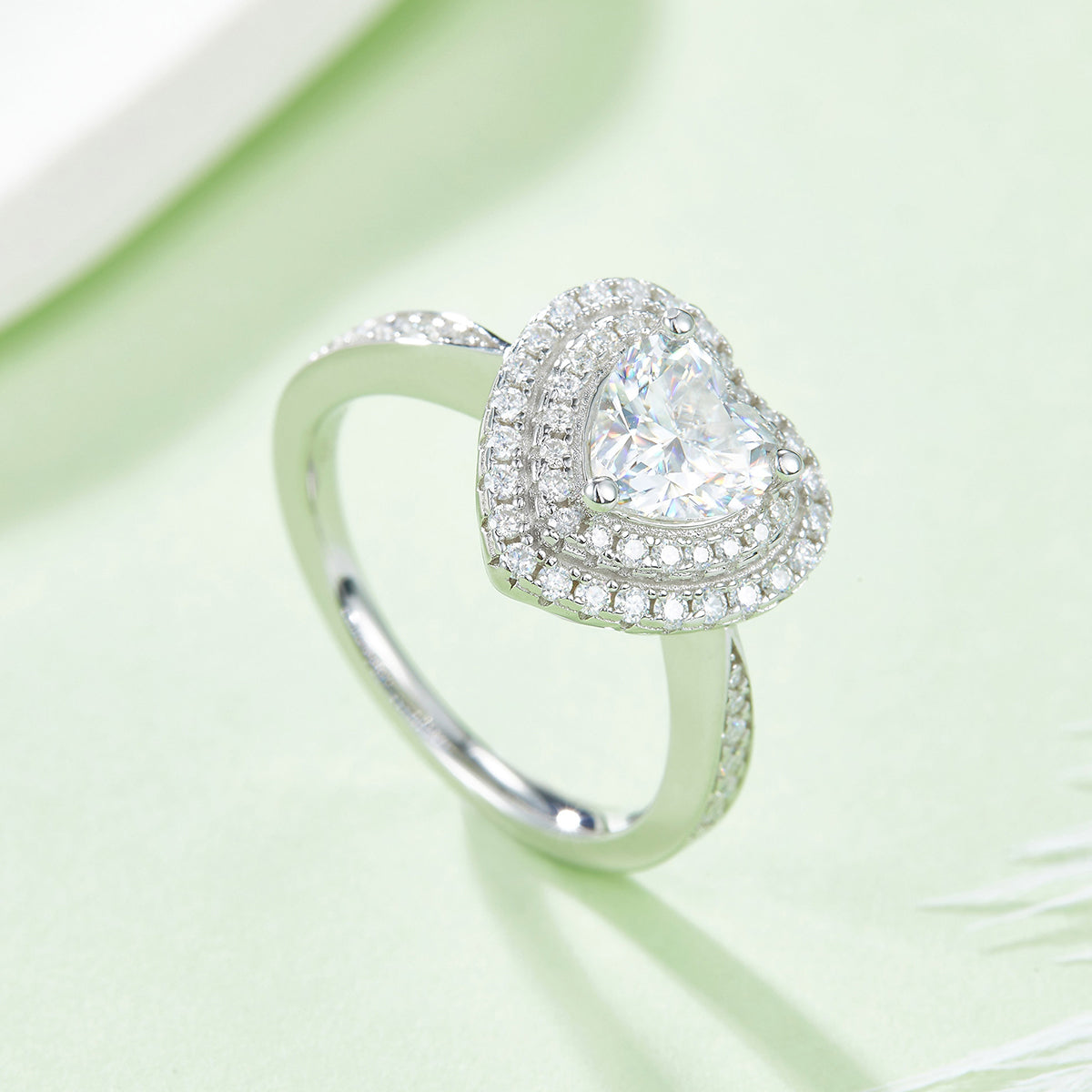 CARISSIE Heart Moissanite Halo Engagement Ring, lifestyle shot on a soft green background showing the ring's sparkle